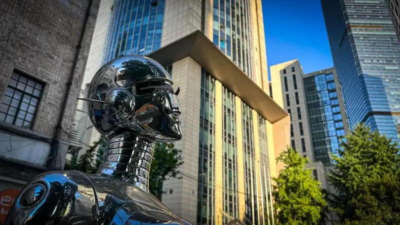 A metallic robot statue stands against modern skyscrapers under a clear sky.